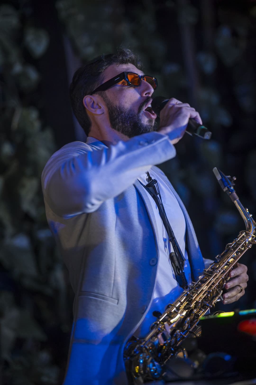 Bruno Daner cantando ao microfone com o saxofone em mãos, sob luz azul